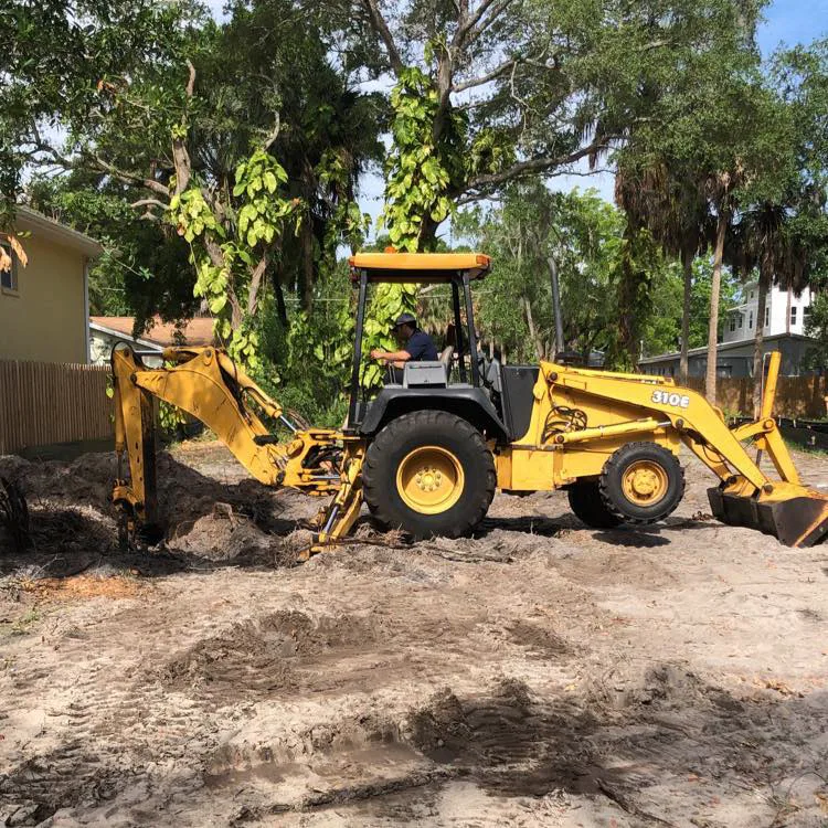 Backhoe digging on a sandy residential lot under mature oaks